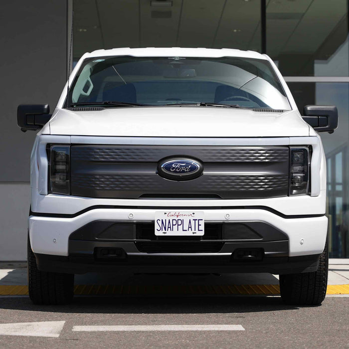 White Ford truck with a custom grille and license plate reading SNAPPLATE, parked in front of a building.
