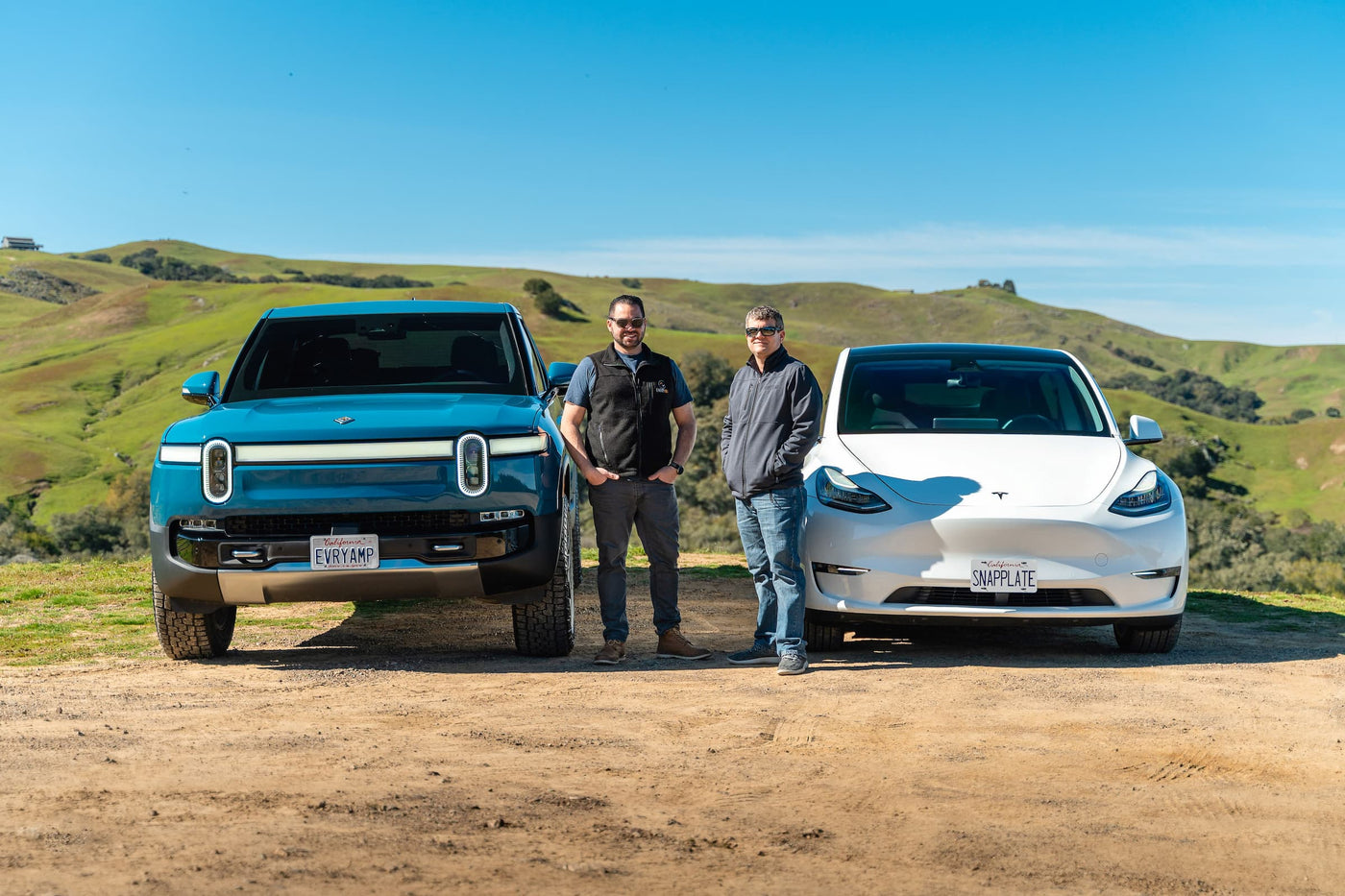 Two individuals stand between a blue truck and a white car on a dirt road with grassy hills under a blue sky.
