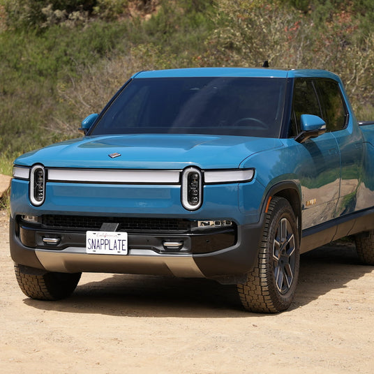 Blue electric pickup truck parked on a dirt road with greenery in the background. License plate reads SNAPPLATE.