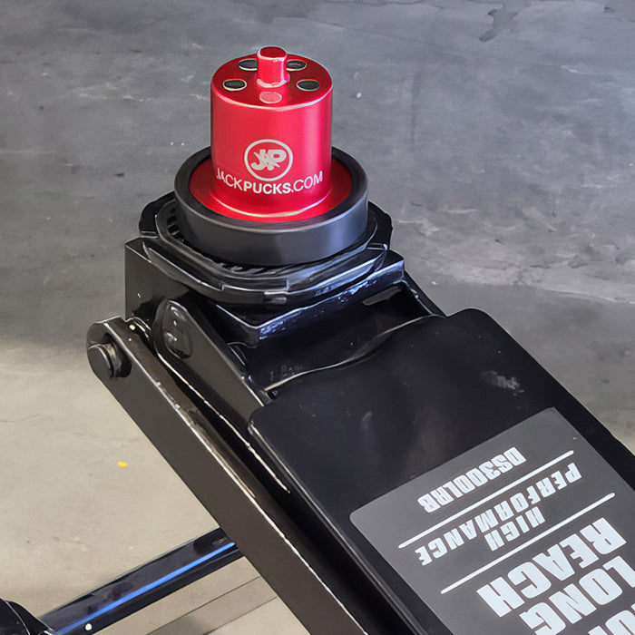 A black car jack with a red adapter labeled JACKPUCKS.COM is positioned on a garage floor.