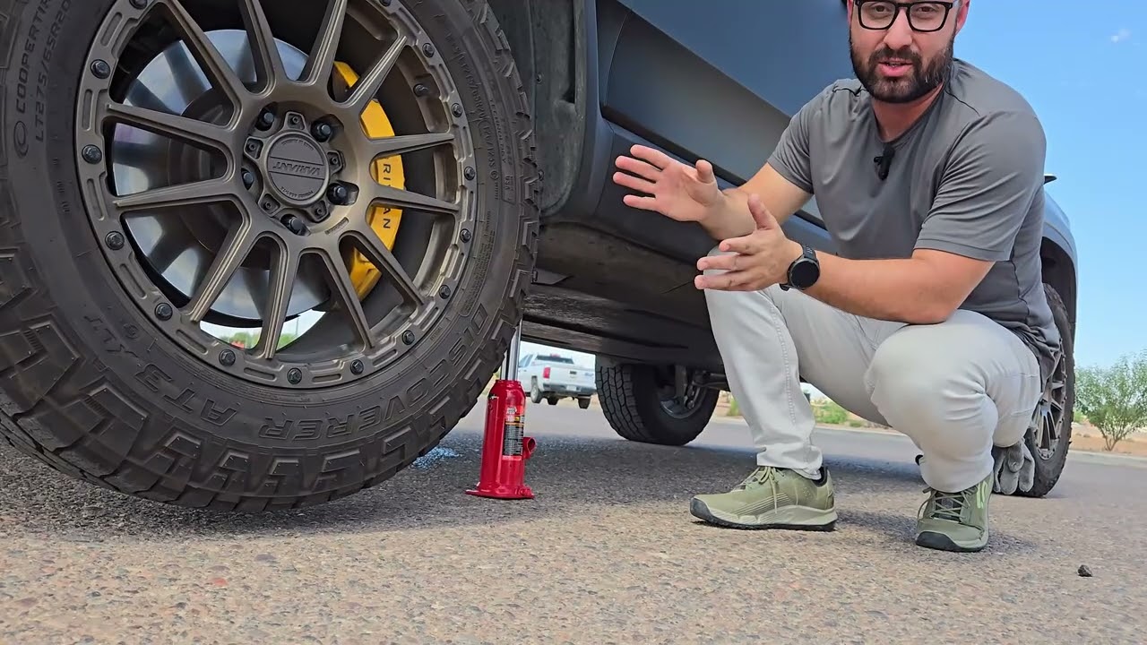 A person crouches next to a jacked-up car wheel on a road, gesturing and speaking. A red bottle jack is visible beneath the car.