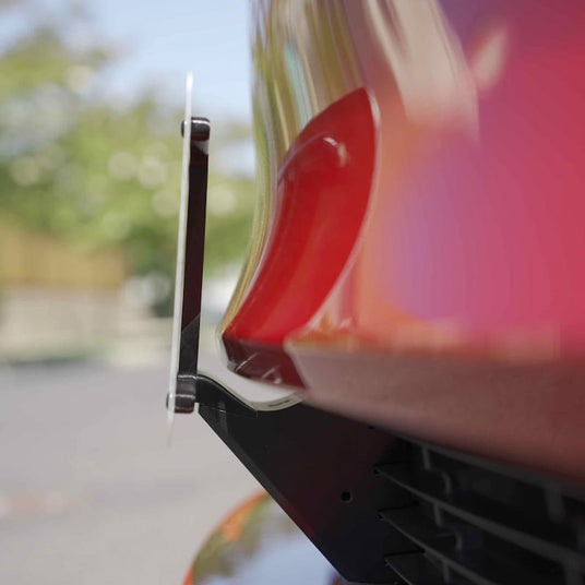 Close-up of a red cars front bumper with a license plate bracket attached, set against a blurred outdoor background.