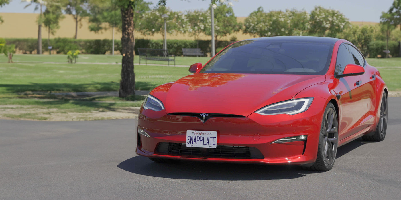 Red Tesla Model S with a SNAPPLATE license plate, parked on a paved road in a park setting with trees and grass in the background.
