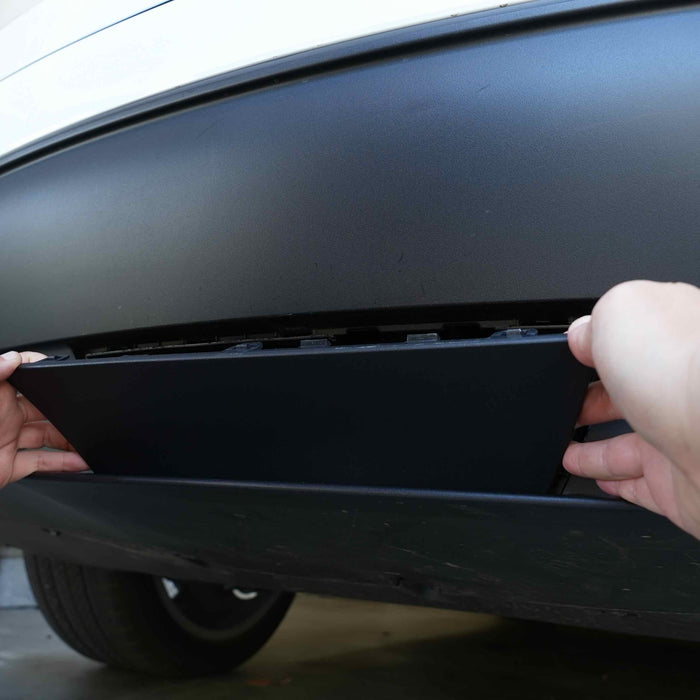 Hands installing a rectangular panel on the underside of a vehicle, below the bumper.
