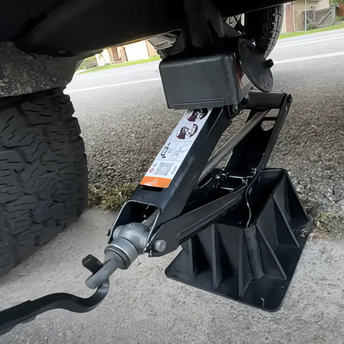A car is raised on the roadside using a black scissor jack and a plastic support stand. The jack is positioned under the vehicle with a handle attached.