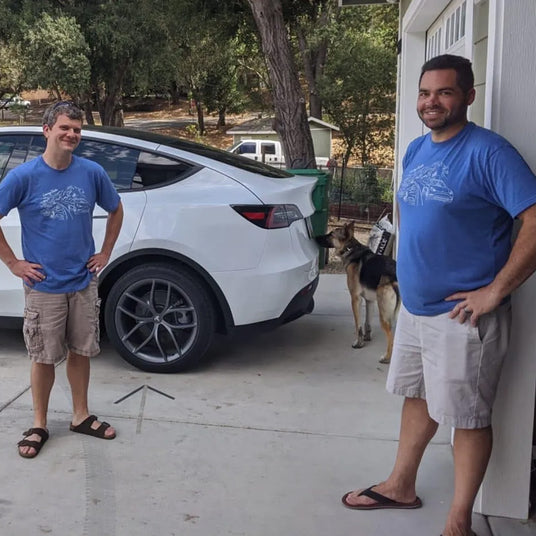 Two men wearing matching blue shirts and khaki shorts stand smiling near a white car in a driveway. A dog stands nearby, partially behind the car.
