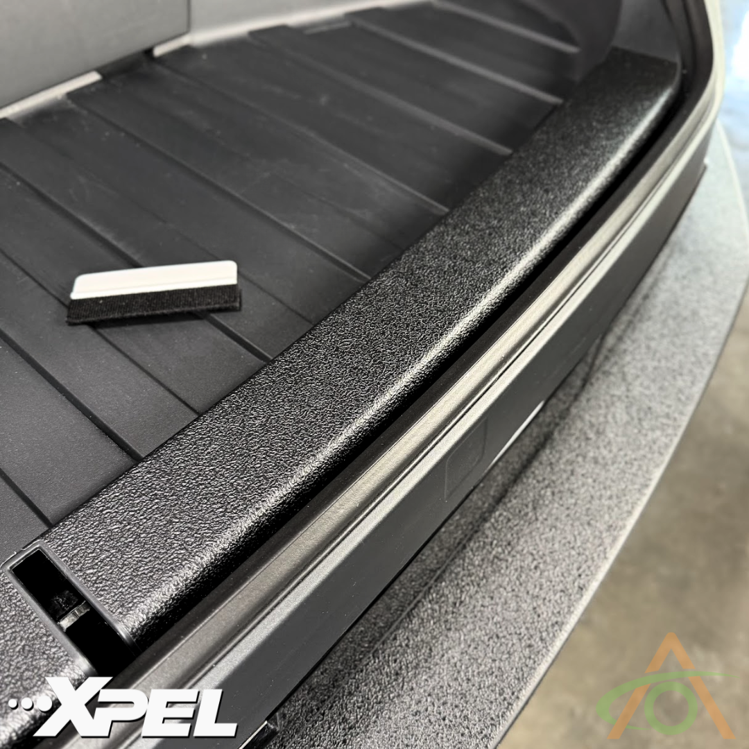 Close-up shot of Abstract Oceans Ultra-Tough Xpel Armor Frunk Protection for Cybertruck, in black textured film being partially installed on a smooth surface, with a squeegee nearby—perfect for protecting your Cybertrucks plastic trim.