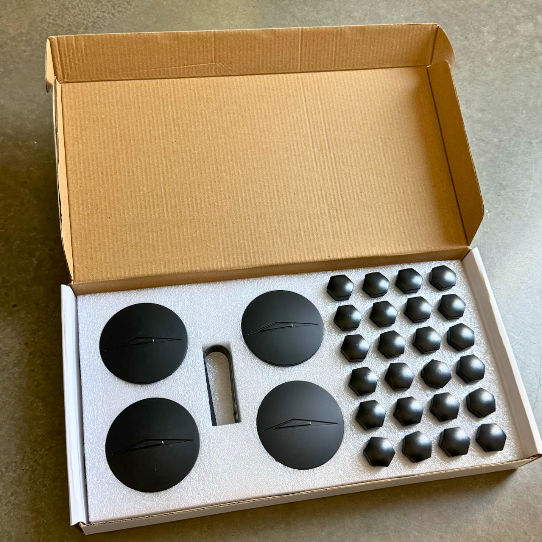 An open cardboard box displays black circular and hexagonal parts, neatly arranged in white foam on a gray surface, showcasing the precision of Abstract Oceans Wheel Center Caps & Lug Nut Covers for Cybertruck.