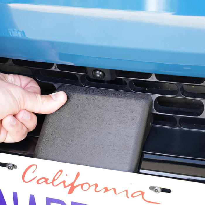 A hand pulls a small rectangular device from the grille of a blue vehicle, located above the California license plate.