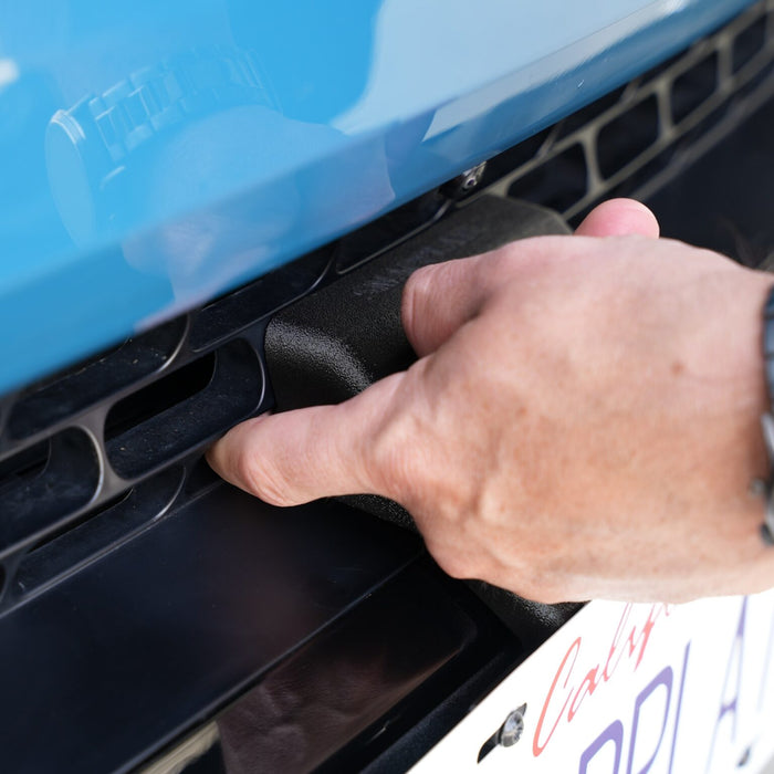 A hand reaches under the front of a blue car, touching a black component near the license plate.