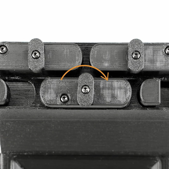 Close-up of a black mechanical component with multiple linked sections fastened by screws. An orange arrow highlights one of the sections.