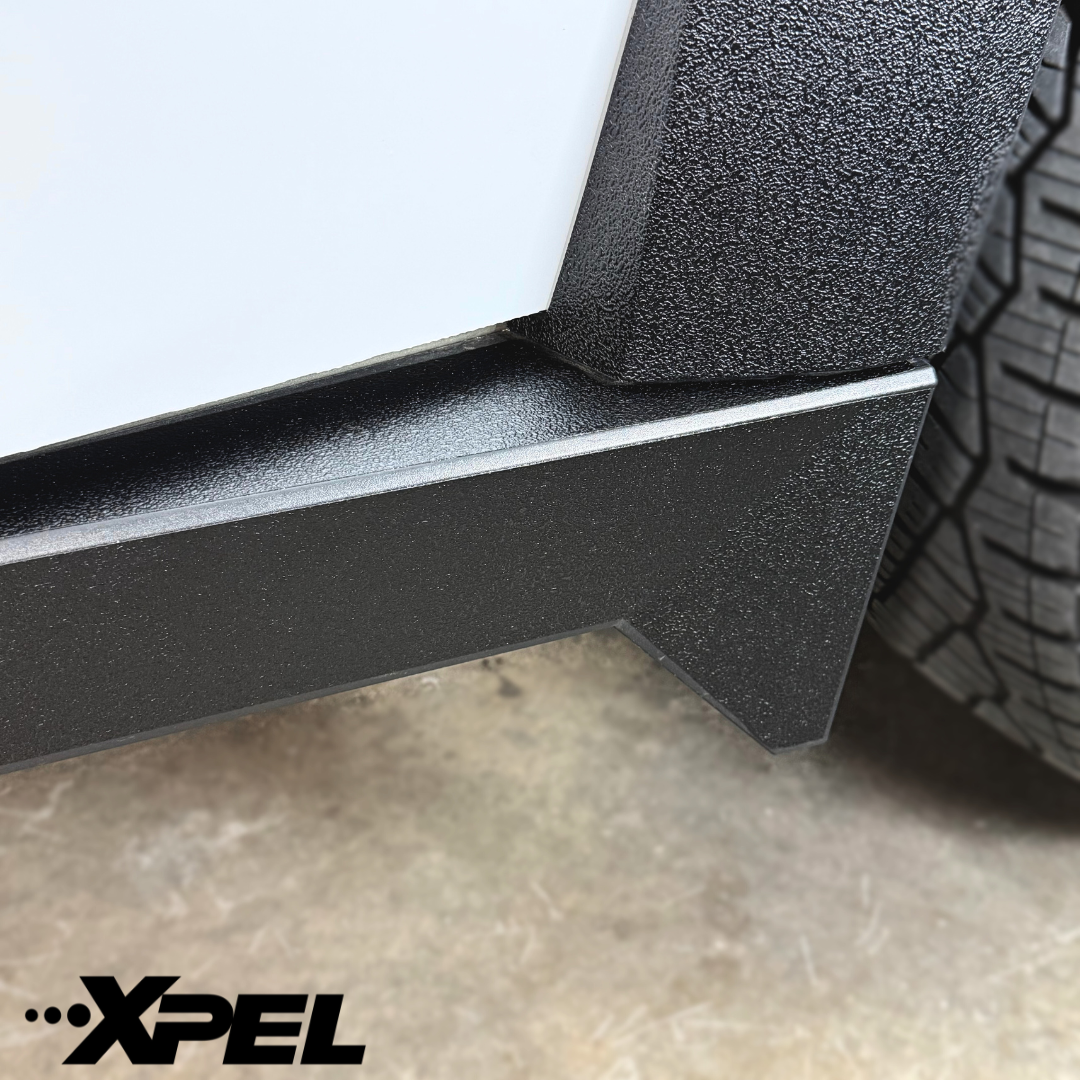 Close-up of a Cybertrucks textured and smooth black trim near the tire on concrete, enhanced with Ultra-Tough Xpel Armor Rocker Protection from Abstract Ocean. Abstract Ocean logo in the bottom left corner.