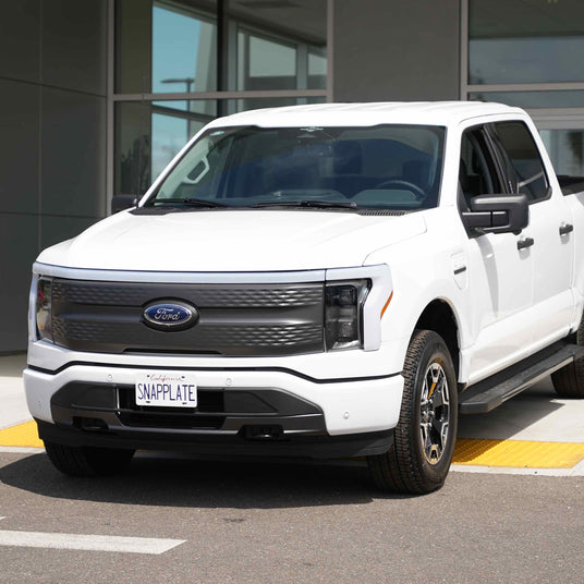 White Ford truck parked outside a building, featuring a license plate with SNAPPLATE on it.