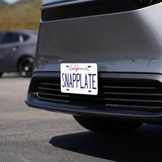 Close-up of a car license plate displaying SNAPPLATE in a parking lot, with another vehicle visible in the background.