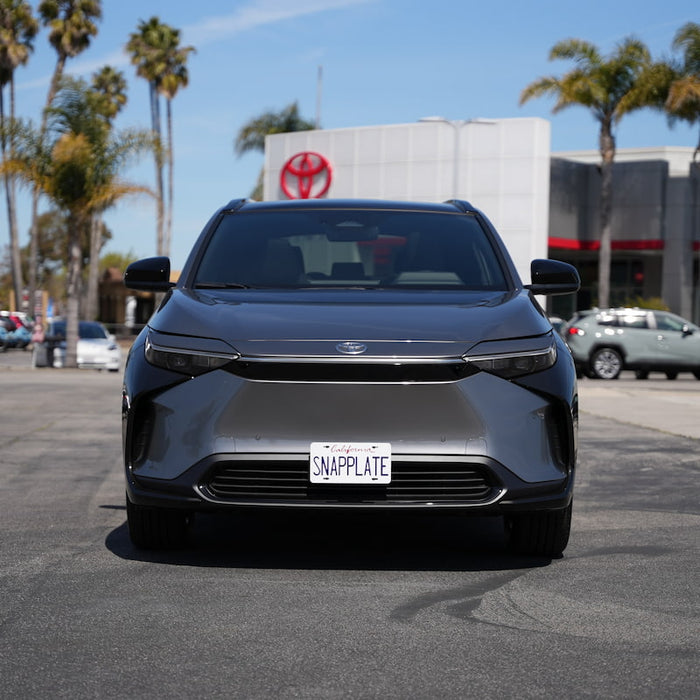 A gray electric SUV is parked in front of a dealership with palm trees and a Toyota logo in the background. The license plate reads SNAPPLATE.