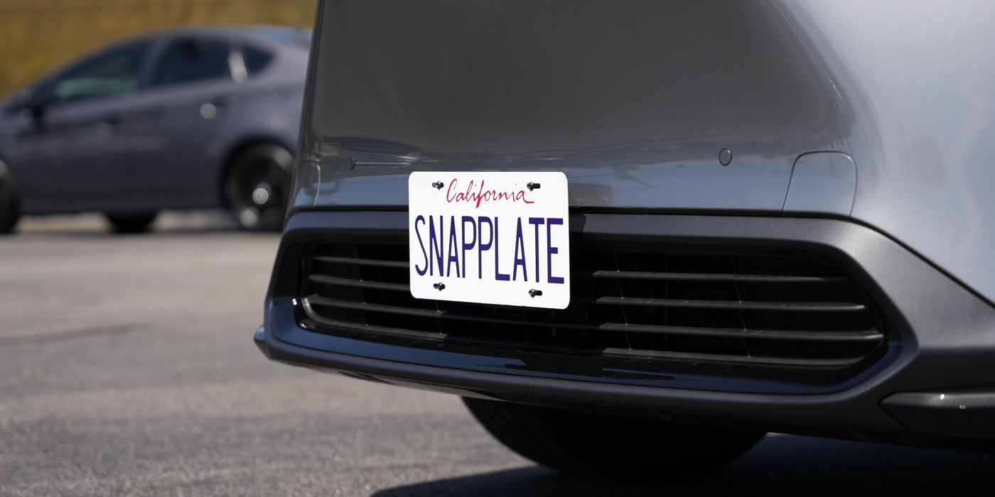 A gray car with a SNAPPLATE license plate holder on the front. Another car is visible in the background on the left.