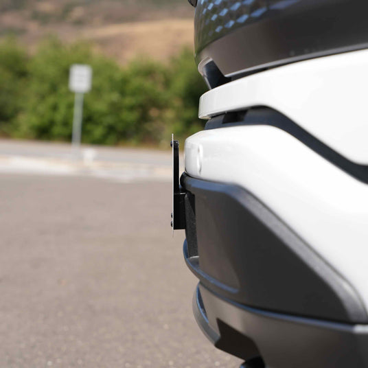 Close-up of a cars rear bumper with a visible parking sensor in an outdoor setting, with blurred greenery in the background.