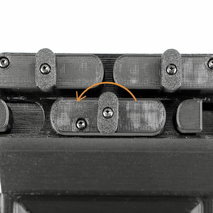 Close-up of a dark mechanical component with several small screws and a central tab indicated by an orange arrow.