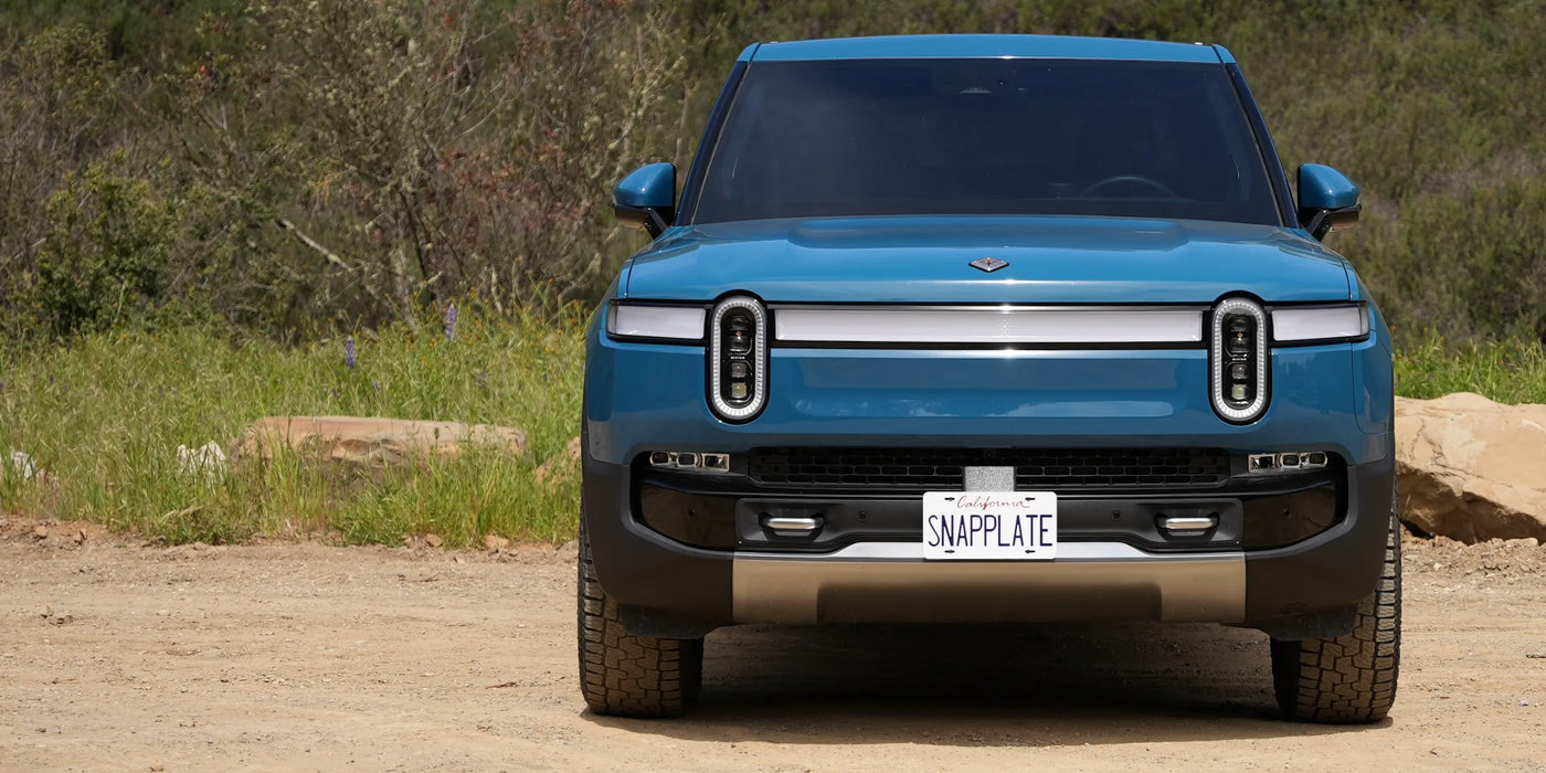 Blue electric SUV parked on a dirt road, featuring circular headlights and a SNAPPLATE license plate, set against a backdrop of greenery and rocks.