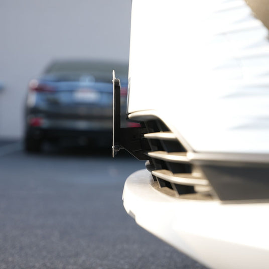 Close-up of a white cars front bumper and license plate holder with a blurred dark car in the background.