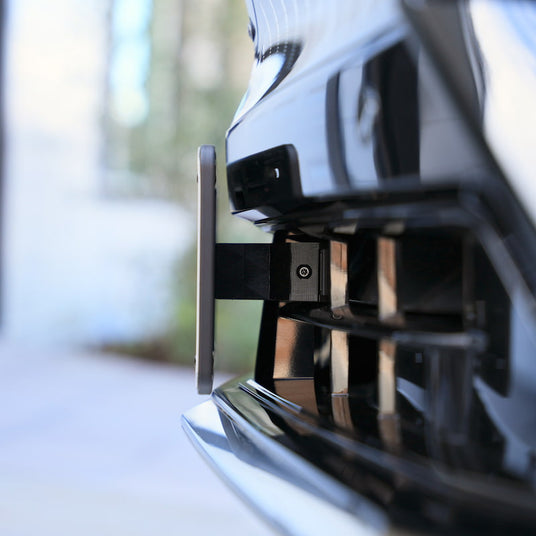Close-up of a cars front bumper showing a rectangular electronic sensor or license plate retracting mechanism, partially extended among the grille vents.