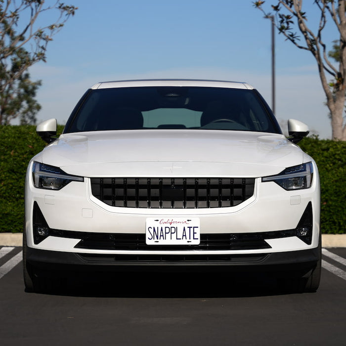 Front view of a white electric car in a parking spot, with a SNAPPLATE license plate, flanked by green hedges and trees.