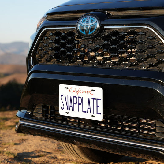 Front view of a black Toyota car with a California license plate reading SNAPPLATE, parked outdoors on a sunny day with a hilly landscape in the background.