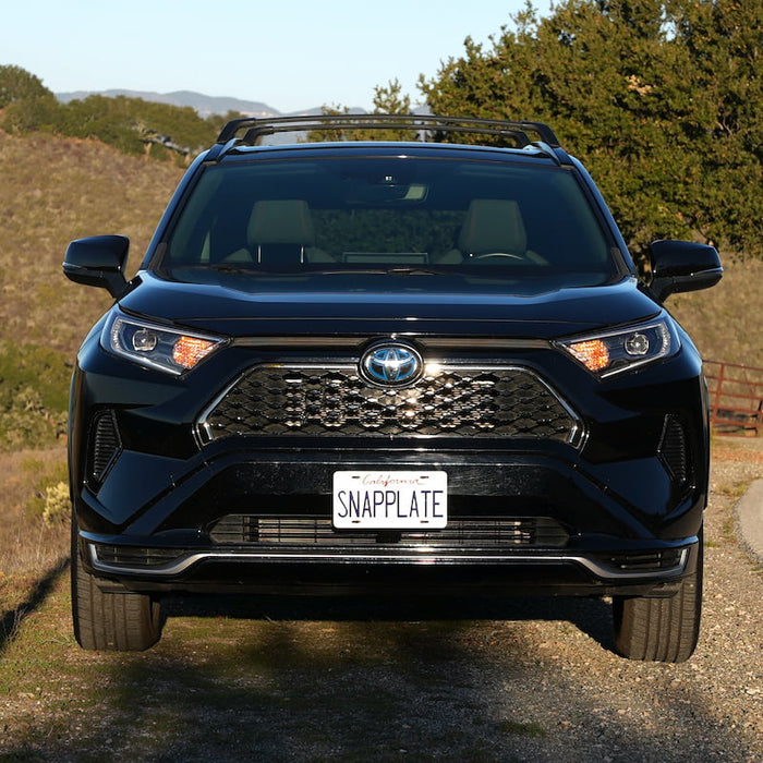 A black Toyota SUV is parked on a dirt path next to a grassy hill, with a personalized license plate reading SNAPPLATE.