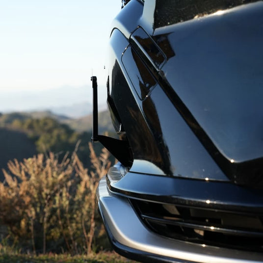 Close-up of a black sports car front end with sleek design details, parked outdoors with blurred greenery and mountains in the background.