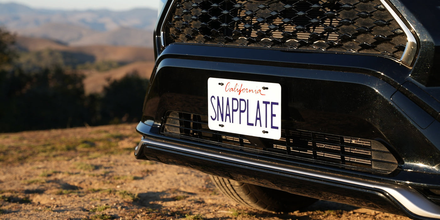Close-up of a black vehicles front with a license plate reading SNAPPLATE in a scenic outdoor setting with mountains in the background.