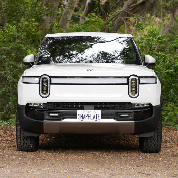 White electric pickup truck with a SNAPPLATE license plate, parked on a dirt path surrounded by green foliage.