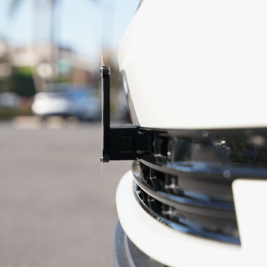 Close-up of a white cars front bumper with a license plate mount, showing a black bracket extending outward. Background is blurred with hints of greenery and other vehicles.