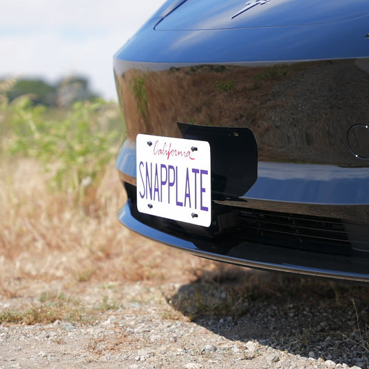 Close-up of a black cars front, featuring a California license plate with the text SNAPPLATE in bold letters, set against a blurred outdoor background with grass and gravel.