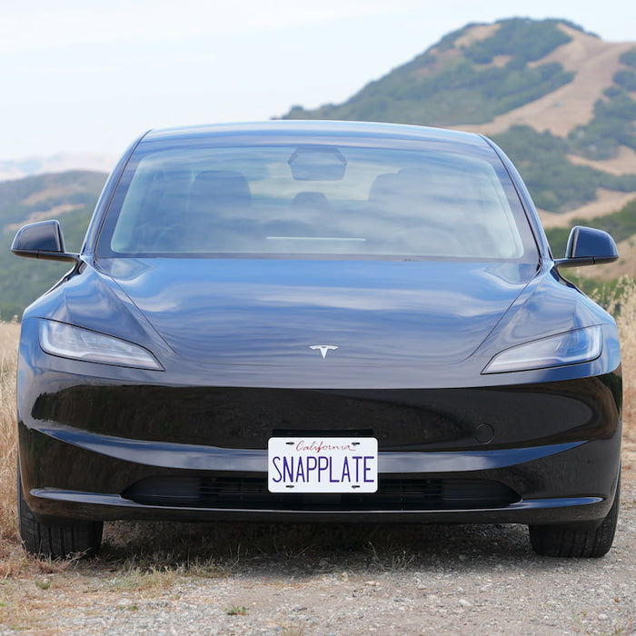 Front view of a black Tesla Model 3 with a Snapplate license plate, parked outdoors with hills in the background.
