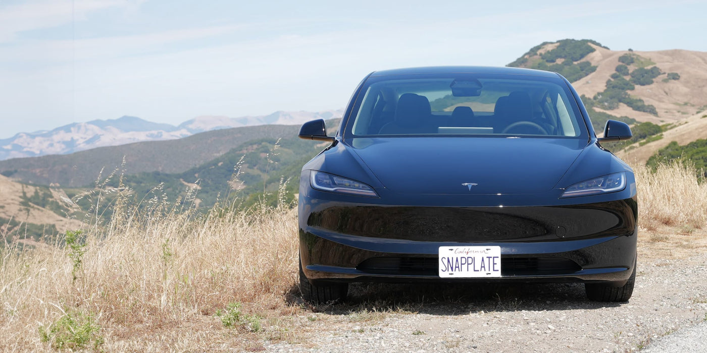 A black Tesla with a customized license plate SNAPPLATE is parked on a grassy roadside with mountains in the background.