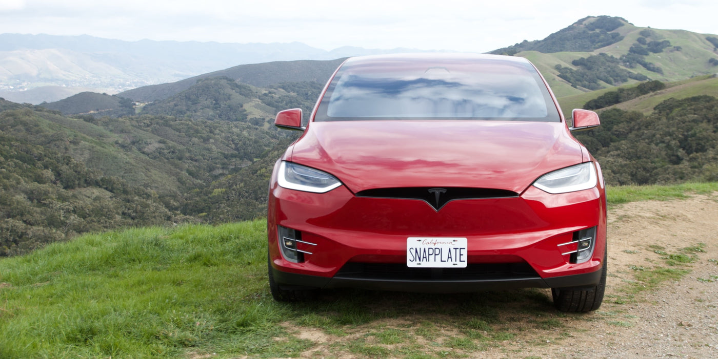 Red Tesla parked on a grassy hill with mountains in the background, featuring a custom license plate reading SWAPPLATE.