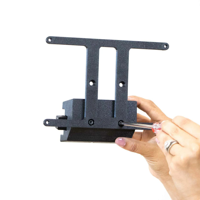 Hands using a screwdriver to assemble or adjust a black metal bracket with two vertical slots and multiple holes, set against a plain white background.