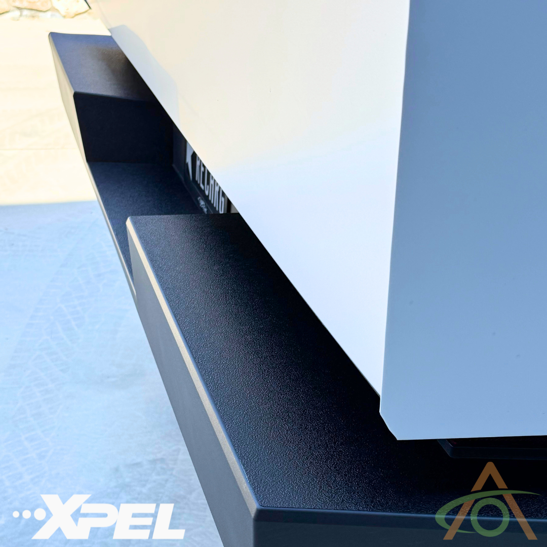Close-up of a white vehicle featuring the Abstract Ocean Ultra-Tough Xpel Armor Rear Bumper Protection for Cybertruck, showcasing a black textured finish with the XPEL Armor logo in the corner.