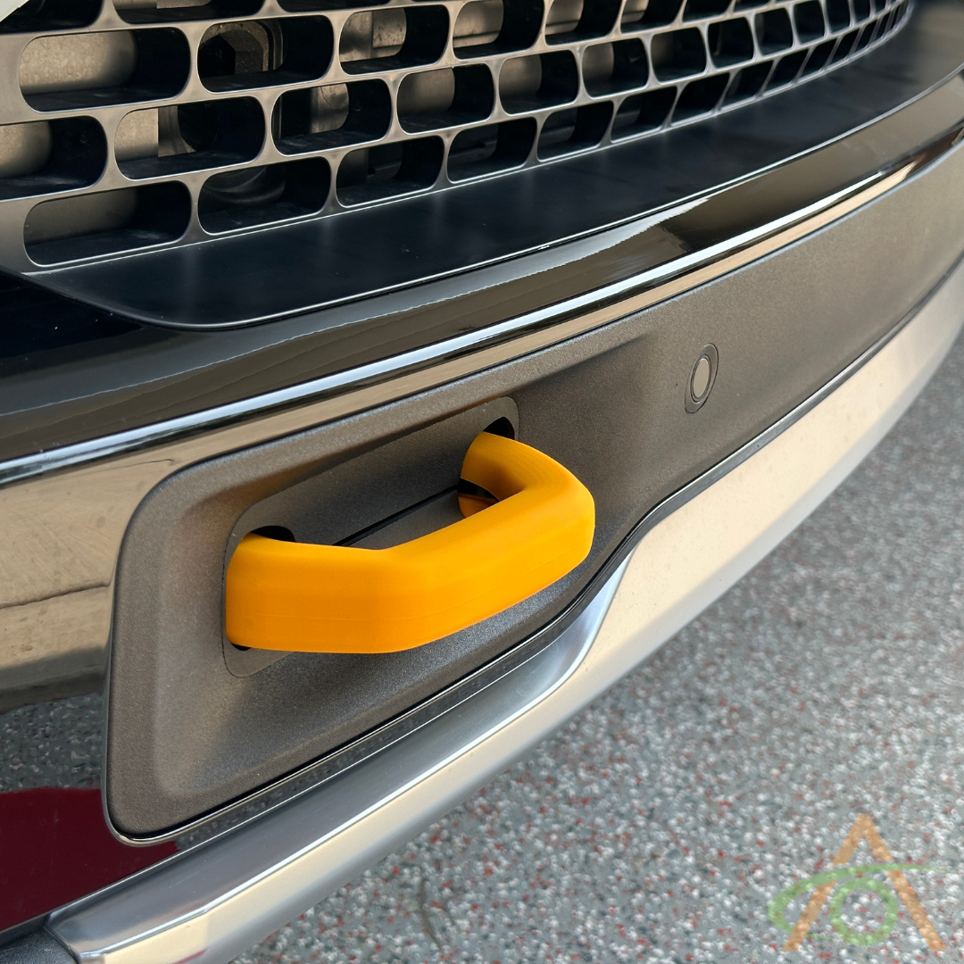 A Rivian Yellow silicone hook cover from Abstract Ocean is attached to the vehicles front bumper with a chrome grille and parking sensor above, enhancing style and function while perfectly complementing other Rivian accessories for an impressive look.