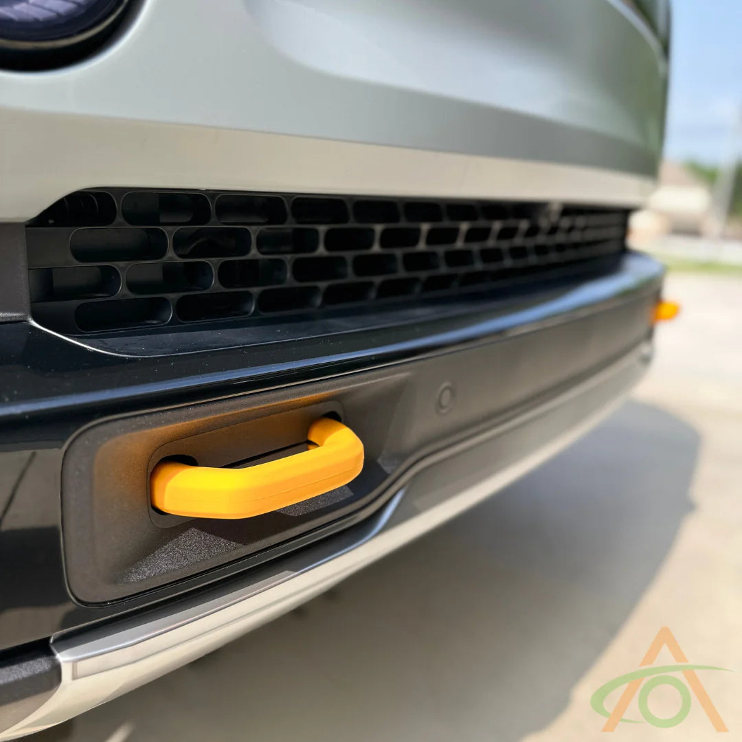 A close-up of a vehicles front bumper features a yellow tow hook with Abstract Ocean Silicone Hook Covers, enhancing the sleek black grille. The blurred ground and building in the background add depth.