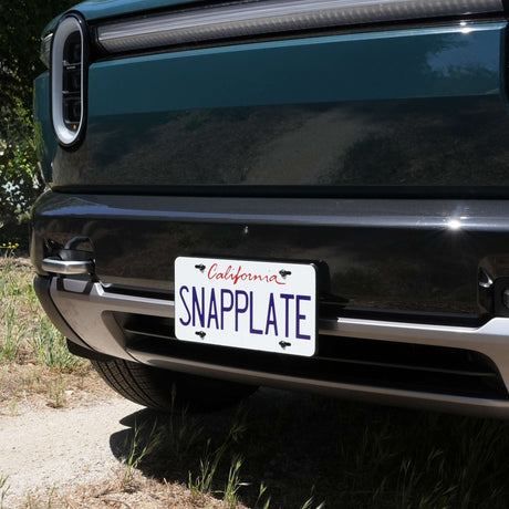 Dark green Rivian R2 with a California license plate on a SnapPlate mount low on the front bumper, the front bumper camera visible just above the plate.