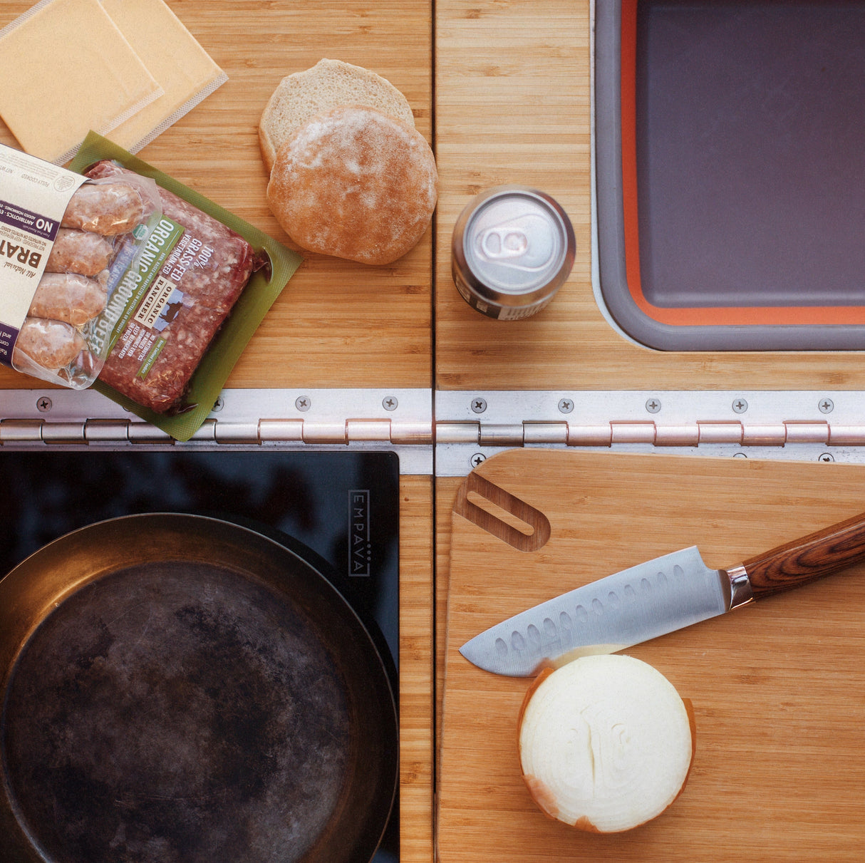 A halved onion, knife, skillet, ground beef, cheese slices, burger buns, and a canned drink rest on the Thunderbolt Adventure Supply Rivian R1T - Overland Kitchen with updated Gear Sled atop a wooden countertop near a sink.