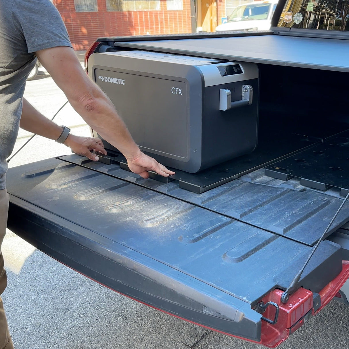 A person slides a Thunderbolt Adventure Supply R1T Bed Glider into the back of a red pickup with the tailgate down, showcasing convenient truck bed storage ideal for organizing camping gear and accessories.