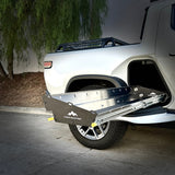 The Range Industries Gear Tunnel Slide 'LITE', compatible with Rivian R1T, in black and silver metal, is extended from the bed of a white pickup truck parked by a curb with trees in the background.