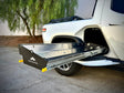 A Range Industries Gear Tunnel Slide ‘LITE’ Compatible With Rivian R1T is extended from the side compartment of a white pickup truck parked on concrete near trees.