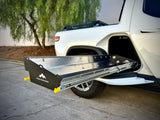 A Range Industries Gear Tunnel Slide ‘LITE’ Compatible With Rivian R1T is extended from the side compartment of a white pickup truck parked on concrete near trees.