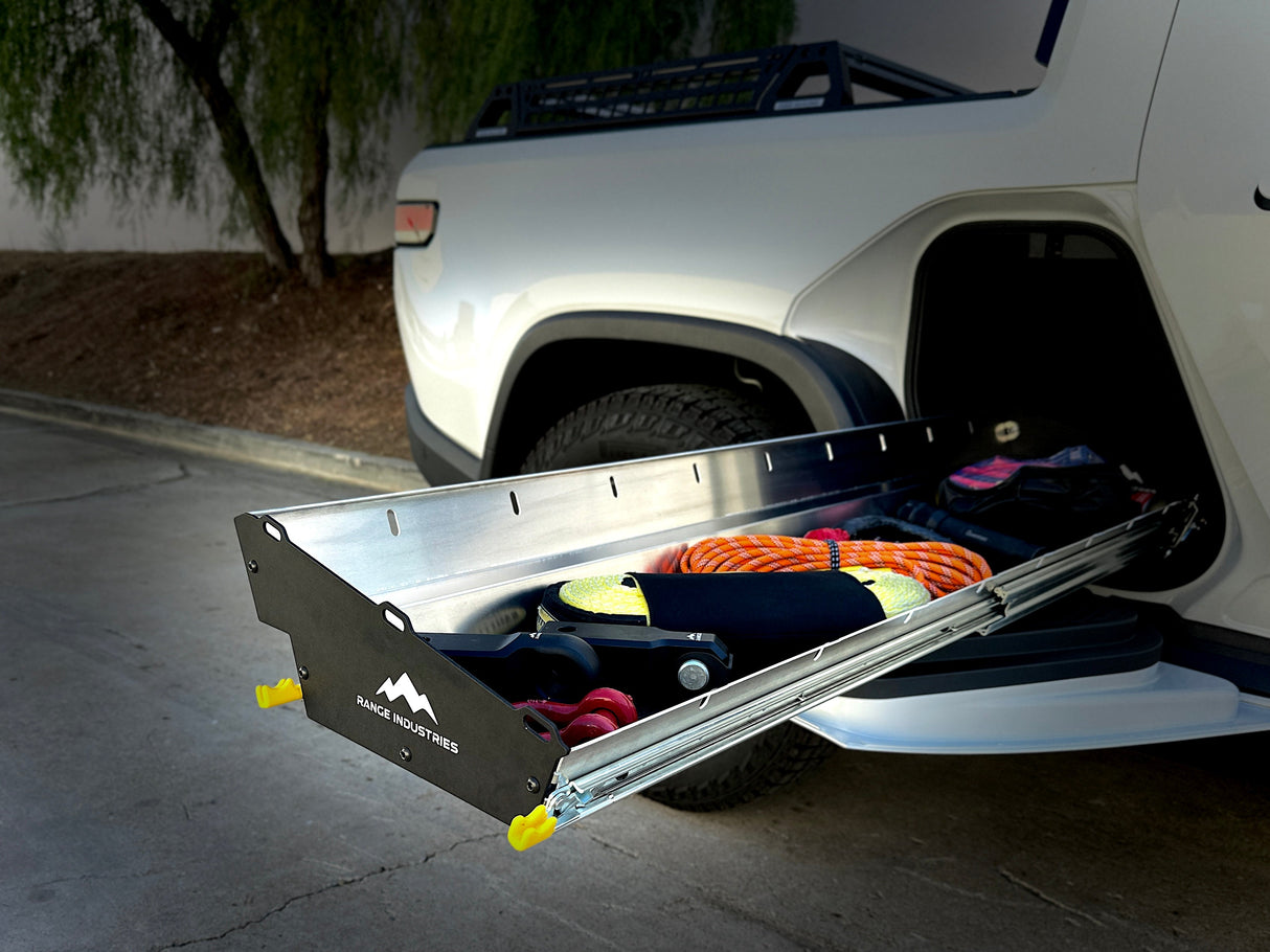 The Range Industries Gear Tunnel Slide 'LITE', compatible with Rivian R1T, extends from a white pickup’s bed, providing practical storage for gear and accessories; a tree and pavement can be seen in the background.