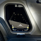 A storage compartment on the vehicle's side holds a black Range Industries Gear Tunnel Slide 'LITE' compatible with Rivian R1T, featuring yellow latches at each corner.