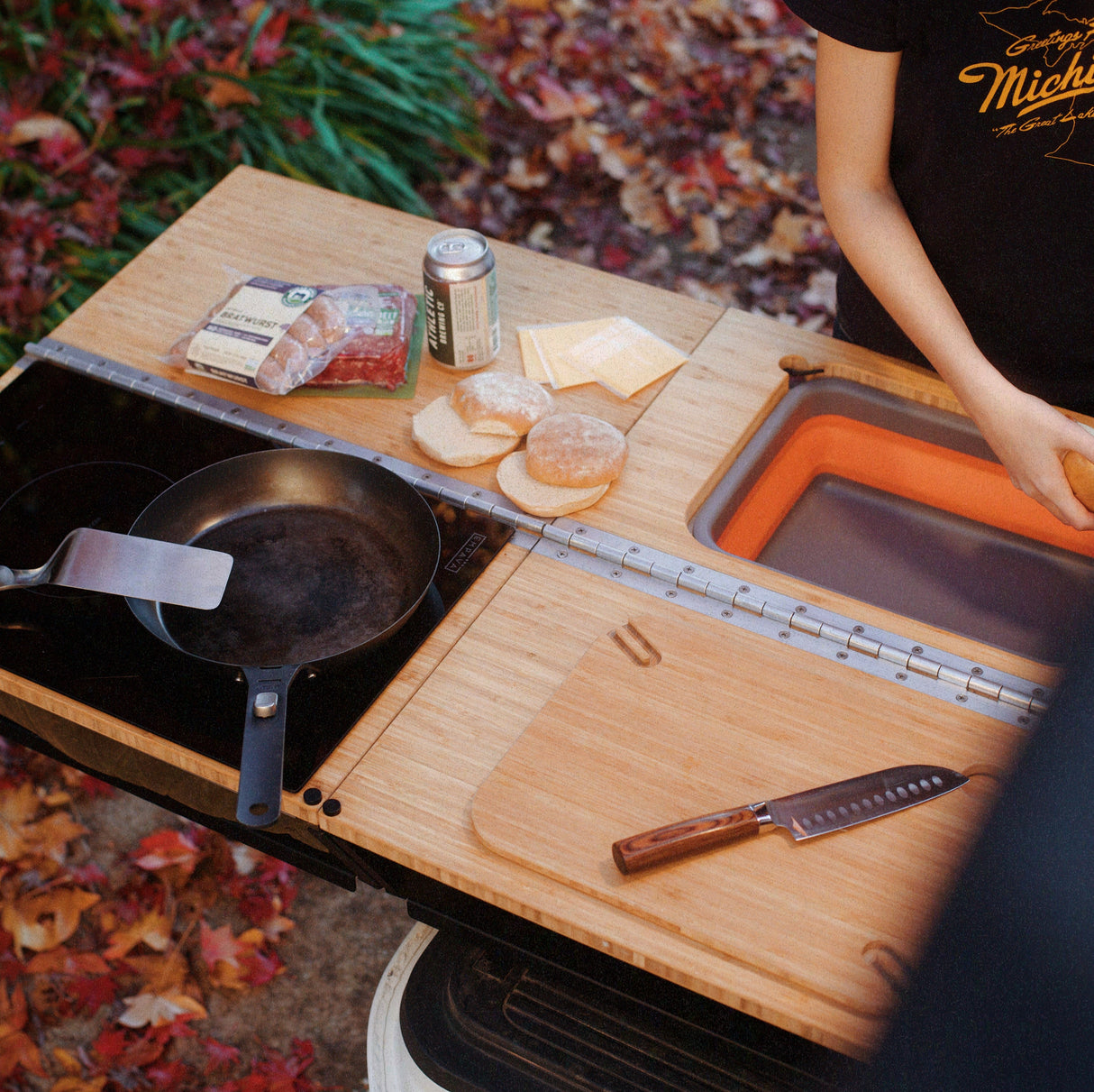 A person stands beside their Thunderbolt Adventure Supply Rivian R1T - Overland Kitchen with updated Gear Sled, featuring a frying pan, spatula, knife, cutting board, burger ingredients, and a drink can.