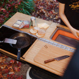 A person stands beside their Thunderbolt Adventure Supply Rivian R1T - Overland Kitchen with updated Gear Sled, featuring a frying pan, spatula, knife, cutting board, burger ingredients, and a drink can.